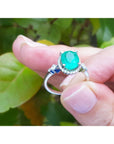 A white gold ring with n oval cut emerald held between fingers against a green leafy background.