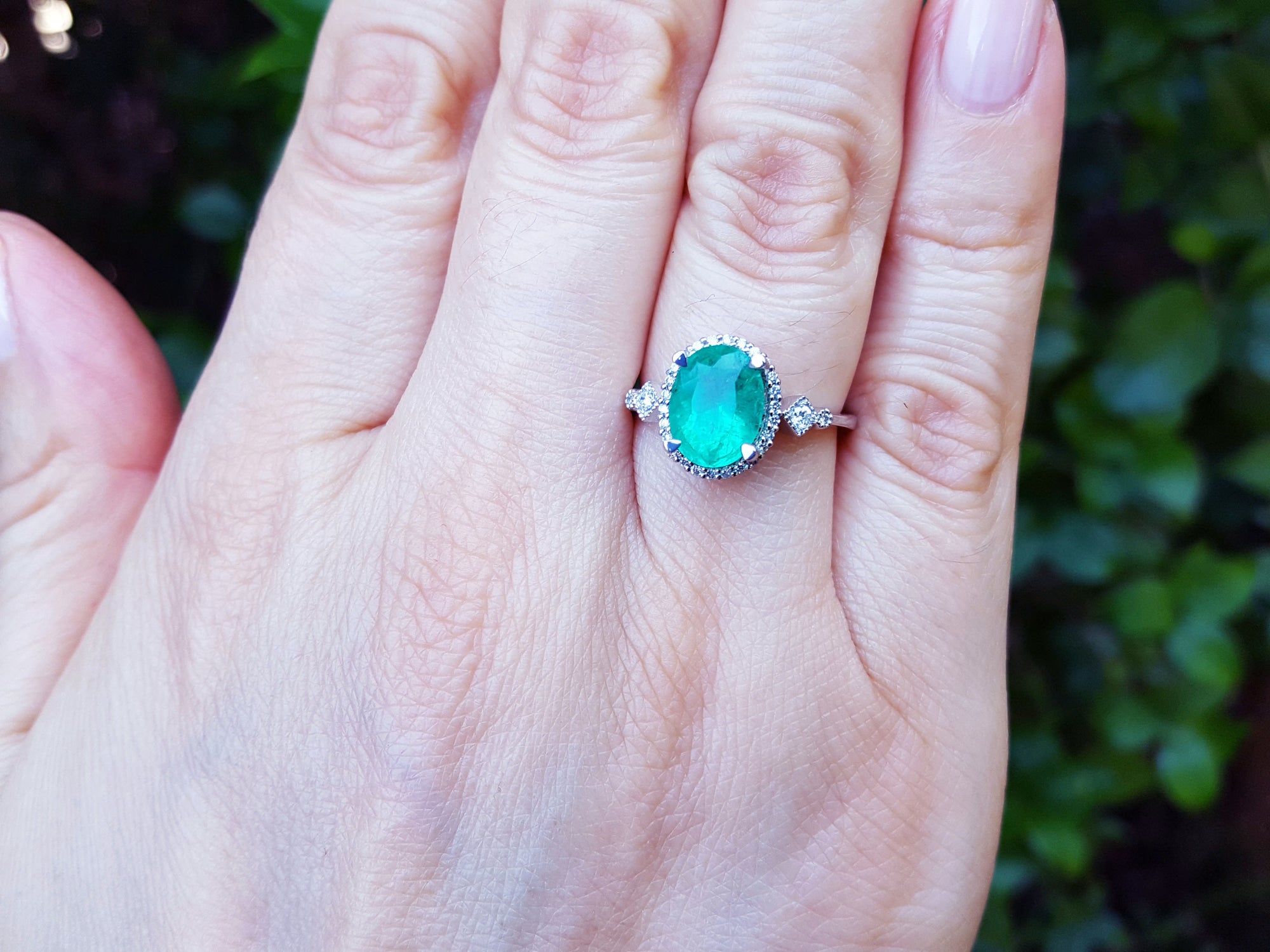 Hand wearing a ring with an oval emerald gemstone against a blurred green background.