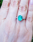 Hand wearing a ring with an oval emerald gemstone against a blurred green background.