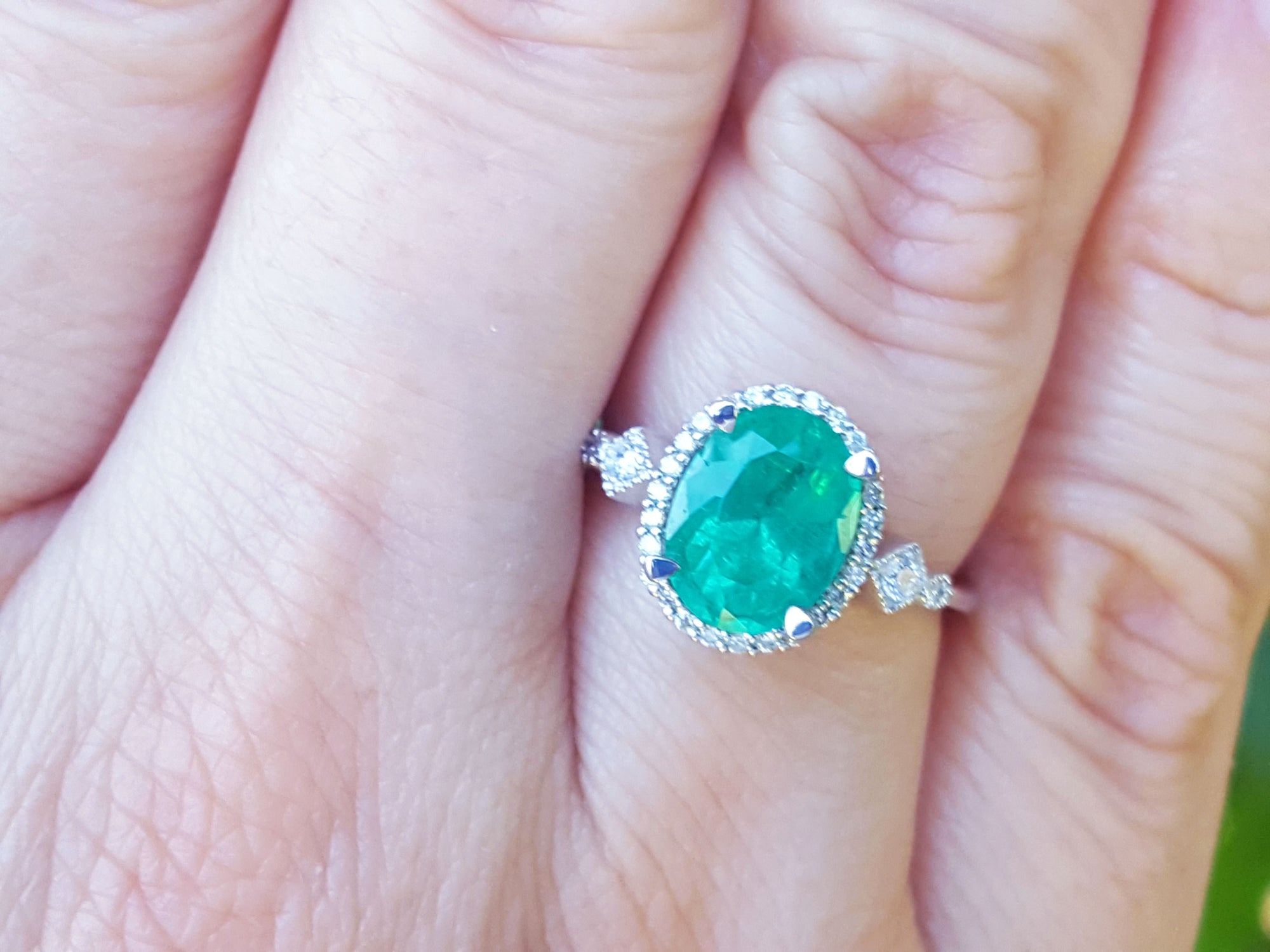 An oval cut emerald ring with accent diamonds on a hand with a blurred background.