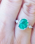 An oval cut emerald ring with accent diamonds on a hand with a blurred background.