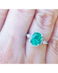 An oval cut emerald ring with accent diamonds on a hand with a blurred background.