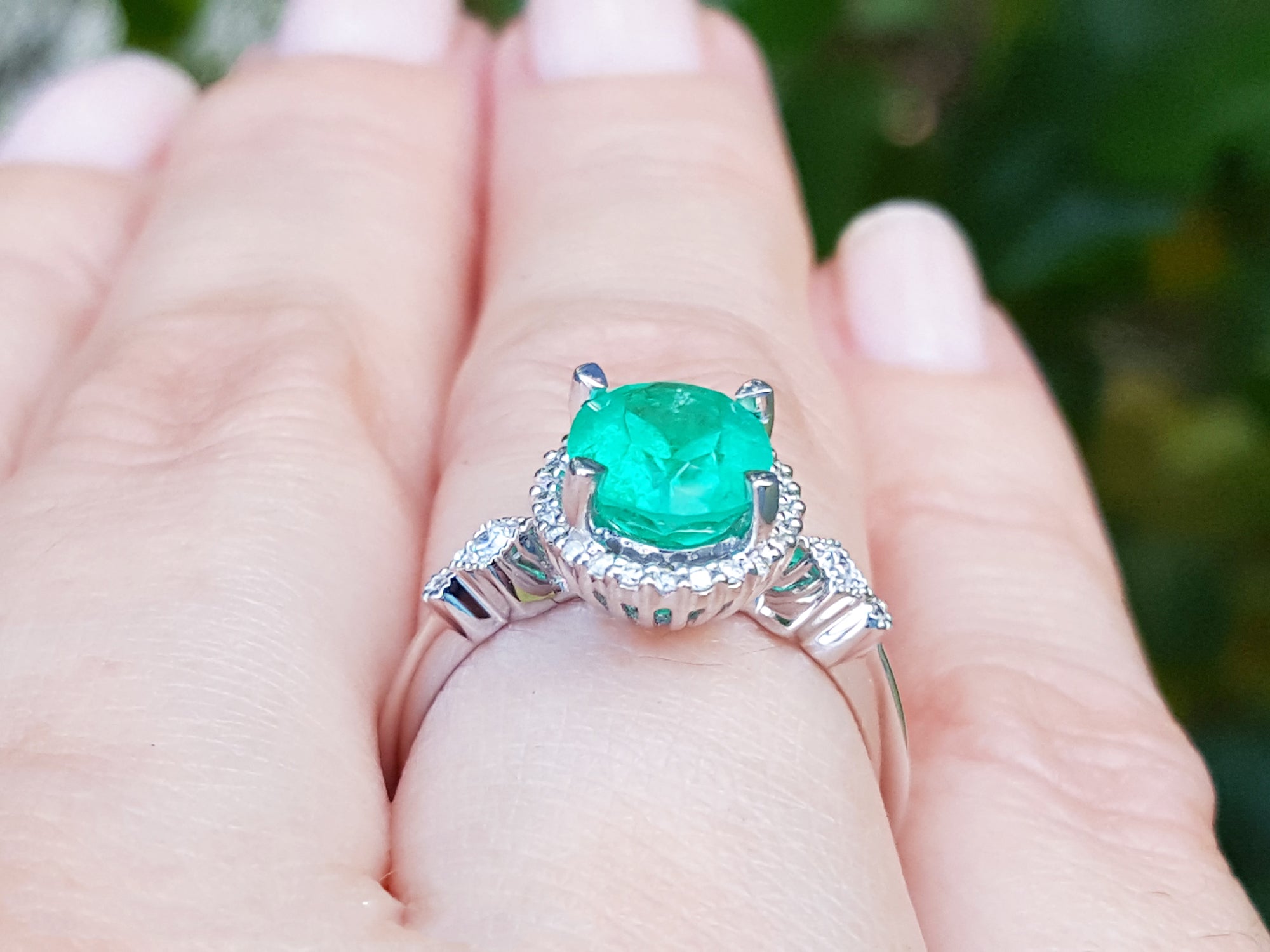 A white gold ring with an emerald and accent diamonds on a hand against a blurred natural background.