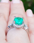 A white gold ring with an emerald and accent diamonds on a hand against a blurred natural background.