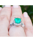 A white gold ring with an emerald and accent diamonds on a hand against a blurred natural background.