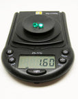 Two oval-cut, deep bluish-green natural emeralds weighing 1.60 cttw on a black digital scale.