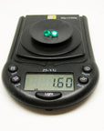 Two oval-cut, deep bluish-green natural emeralds weighing 1.60 cttw on a black digital scale.