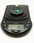 Two oval-cut, deep bluish-green natural emeralds weighing 1.60 cttw on a black digital scale.