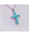 18K white gold cross pendant with round-cut emeralds on a delicate chain, worn on a person.