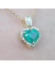 Heart-shaped emerald pendant with diamond accents on a gold chain against a light pink background