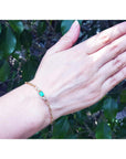 A woman's hand wearing a gold paperclip bracelet with natural emerald against a natural background.