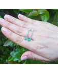 Hand displaying a pear shaped emerald and diamond earrings against a green leafy background