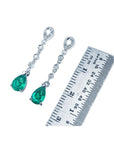 Pair of emerald teardrop earrings with diamond accents next to a ruler for scale on a white background