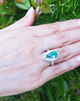 Hand wearing a ring with a pear shaped emerald against a green leafy background.
