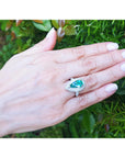 Hand wearing a ring with a pear shaped emerald against a green leafy background.