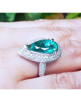 A bridal's pear shaped emerald engagement ring on a person's finger against a blurred natural background.