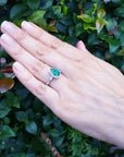 Hand wearing a teardrop emerald ring against a green leafy background.