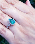 Hand wearing a ring with a pear cut emerald gemstone against a blurred natural background.