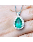 Pear-shaped emerald pendant with halo diamonds in 14K white gold, held by a hand.