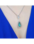Teardrop emerald pendant with double diamond halo on an 18K white gold chain, worn on a blue dress.