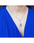 Teardrop emerald pendant with double diamond halo on an 18K white gold chain, worn on a blue top.