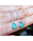 Teardrop-shaped emerald gemstone earrings on a hand with a blurred background