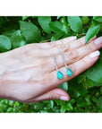 Pear shape 5.05 ct. emerald drop earrings in 18K white gold, held against green leaves.