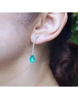 Pear shape 5.05 ct. emerald drop earrings in 18K white gold with diamond accents, worn on an ear.