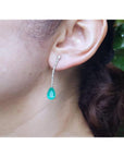 Pear shape 5.05 ct. emerald drop earrings in 18K white gold with diamond accents, worn on an ear.