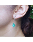 Pear shape 5.05 ct. emerald drop earrings in 18K white gold with diamond accents, worn on an ear.