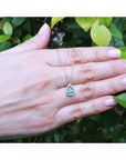 Pear-shaped emerald and diamond halo necklace in 14K yellow gold, held against green leaves.