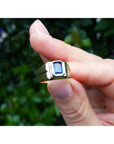 Between fingers holding a ring with a blue sapphire gemstone against a blurred green background