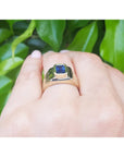 Gold solitaire ring with a blue sapphire gemstone on a hand against a green leafy background