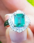 Emerald ring with diamond halo held in a hand against a blurred natural background.