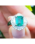 Emerald ring with diamond halo held in a hand against a blurred natural background.