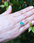 Hand wearing a ring with colombian emerald against a leafy background.