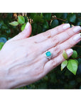 Hand wearing a ring with colombian emerald against a leafy background.