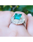 Ring with a Muzo emerald and diamond accents on a finger against a blurred natural background.