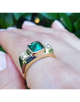 A men's gold ring with emerald and diamond on a hand against a blurred green background.