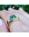 A men's gold ring with emerald and diamond on a hand against a blurred green background.