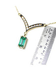 14K yellow gold necklace with a baguette-cut Colombian emerald pendant dangling from a diamond-accented curved V, shown next to a ruler for scale.