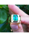 Gold solitaire ring with a rectangular emerald cut held between fingers against a green leafy background.
