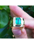Gold solitaire ring with a rectangular emerald cut held between fingers against a green leafy background.