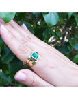 Hand wearing a ring with a genuine emerald gemstone against a leafy background.