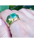 A 18k yellow gold ring with a Muzo colombian emerald on a hand against a natural background.