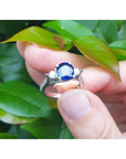 white gold ring with a blue sapphire gemstone held between fingers against a green leafy background