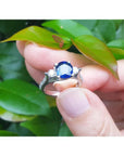 14K white gold ring with a deep blue oval sapphire and two side diamonds, held against green leaves.