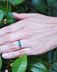 A hand wearing a three stone emerald ring on a leafy background.
