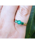 Round cut three-stone emerald ring with prong-set stones in white and yellow gold on a finger.