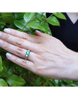 Hand wearing a real Colombian emerald ring against a green leafy background.